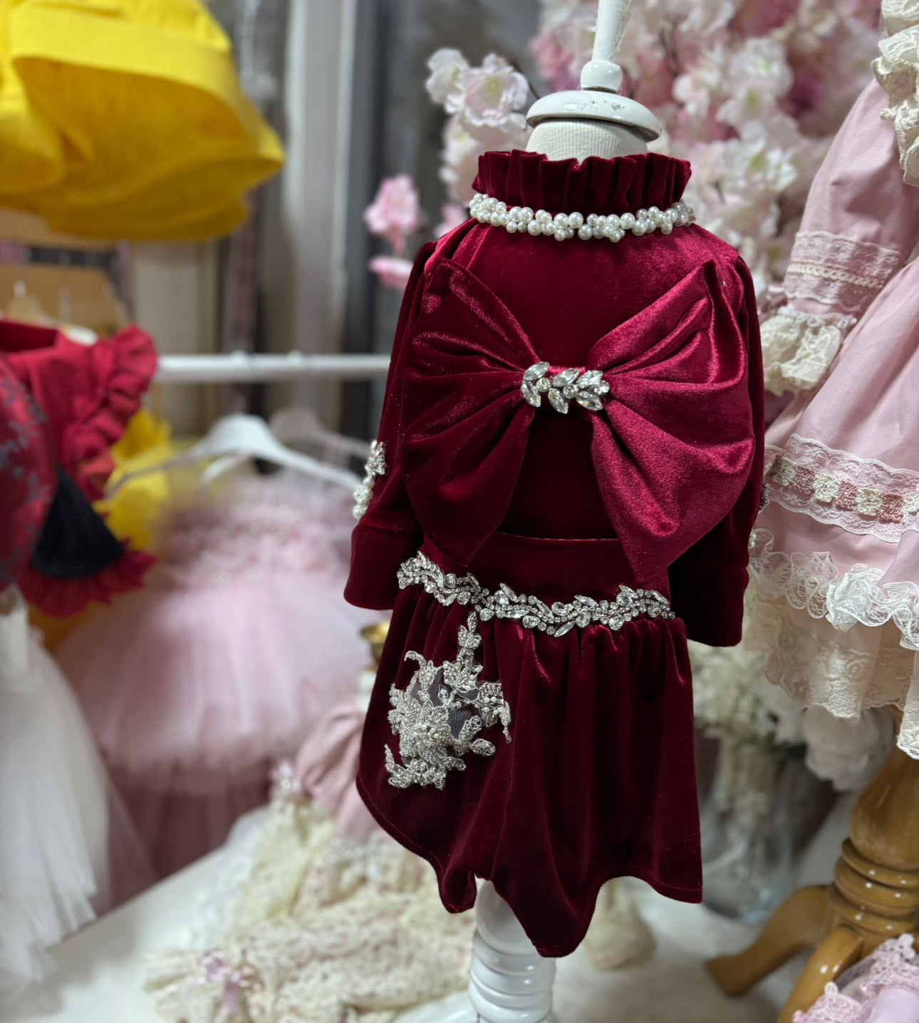 Red velvet dress with decorative bow and embellishments on a mannequin against a blurred background of other dresses.